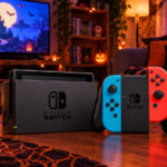 Une Nintendo Switch avec sa base et ses manettes Joy-Con bleue et rouge, posée sur une table basse dans un salon décoré pour Halloween, entre citrouilles, bougies et guirlandes lumineuses